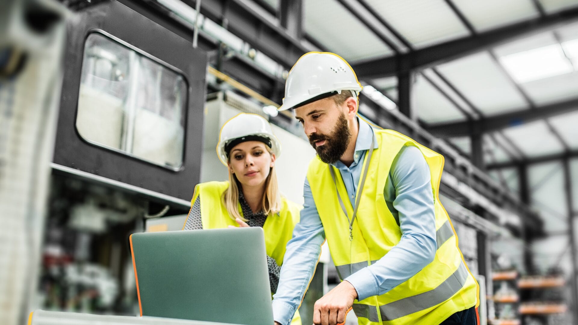 Two manufacturing professionals wearing safety vests and hard hats review information on a laptop inside a factory, representing digital and omnichannel buying experiences in B2B manufacturing.